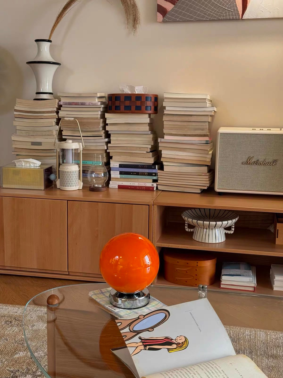 Stacks of books on a wooden shelf with an orange spherical object and a magazine on a glass table.