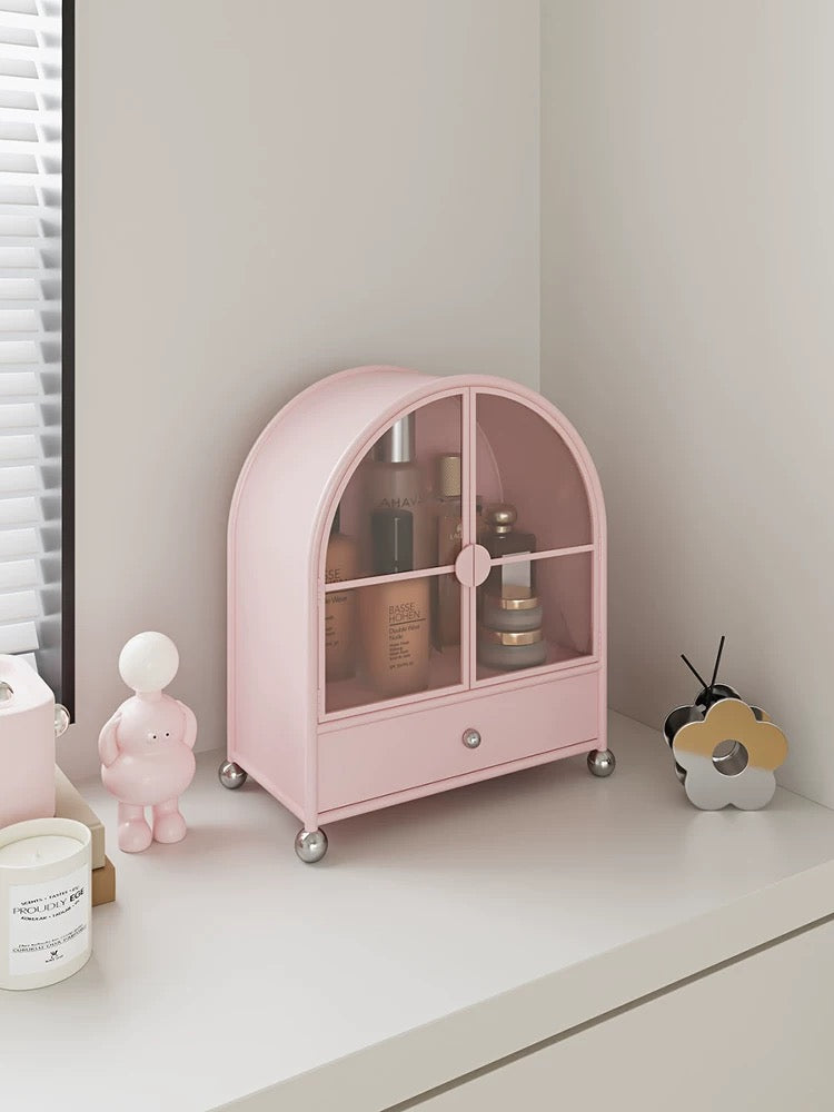 A pink makeup cabinet stands at an angle, showing its curved top, glass doors and silver ball feet.