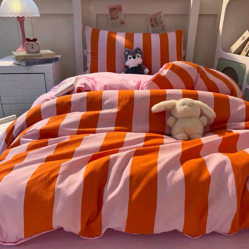 A cozy bed styled with orange and white striped bedding, including a duvet cover and pillowcase, with a pink pillow. A small plush bunny toy rests on the bed, and a small cat toy is on the headboard.