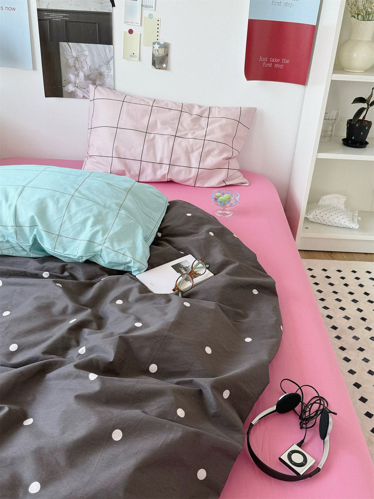 Pink polka dot bedding with blue and pink checked pillows above a small dotted cover and plain pink sheet.