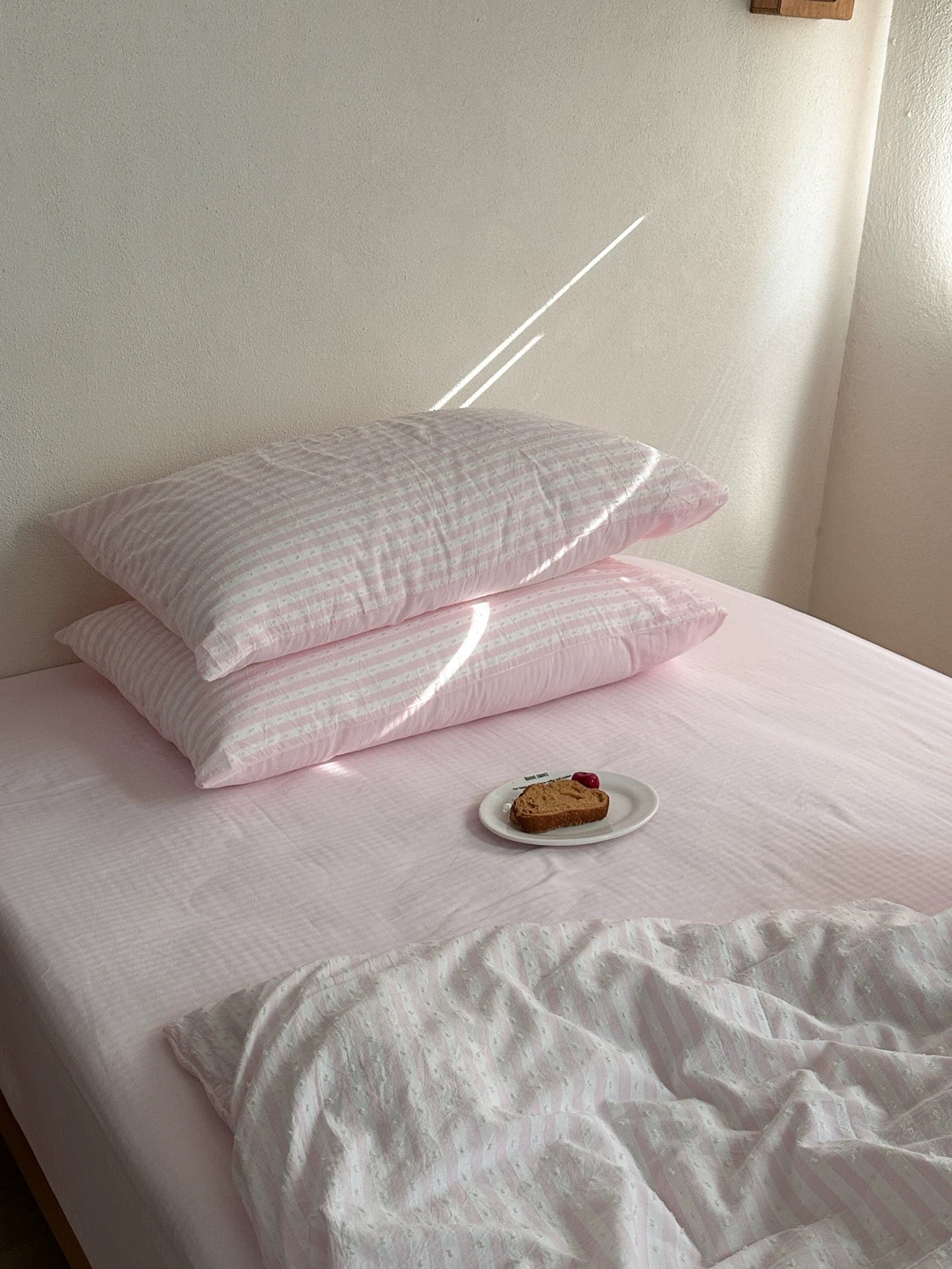 A bed with light pink striped bedding, featuring two stacked pillows, a white duvet, and a small plate with a slice of cake in a softly lit room.