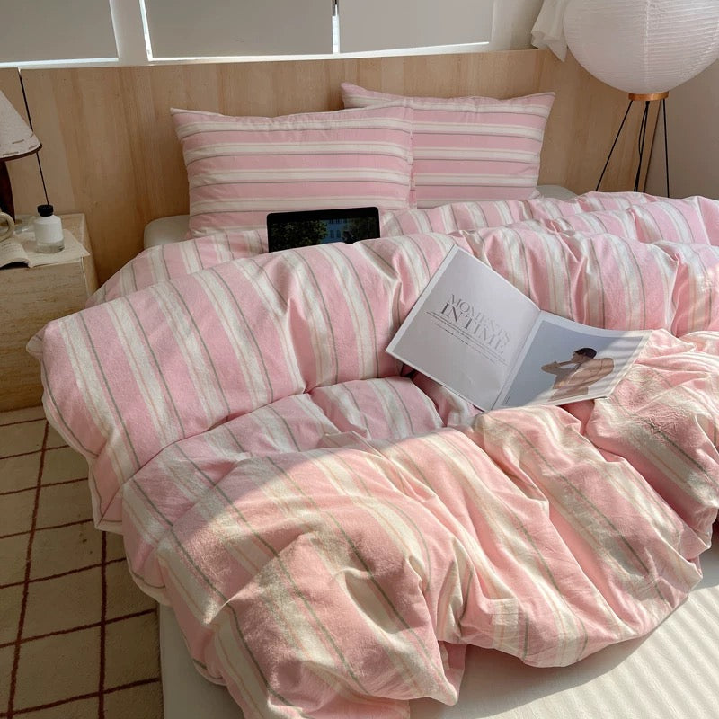 Pink and white striped bedding set on a bed with a side table and lamp in the background.