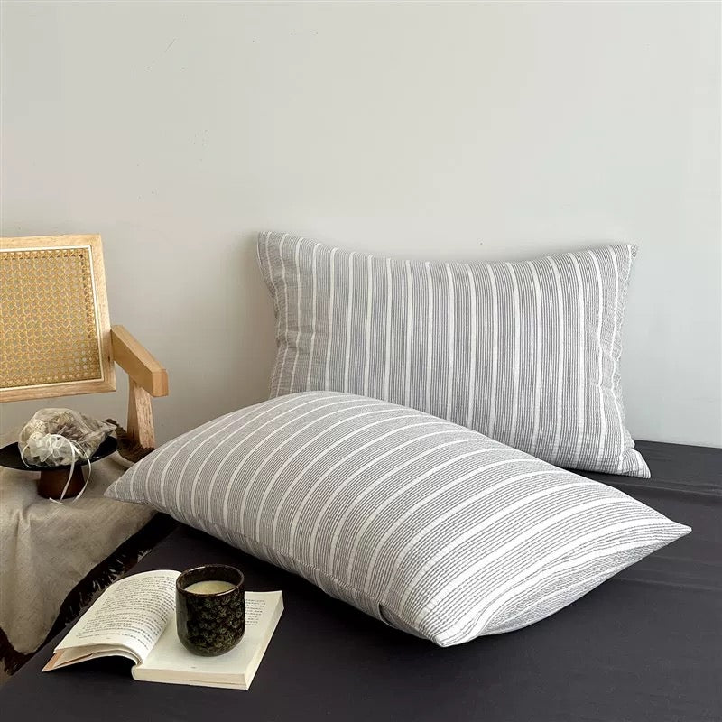 A bed made with a light gray and white striped bedding set, including a duvet cover and two matching pillowcases, styled in a minimalist bedroom setting.
