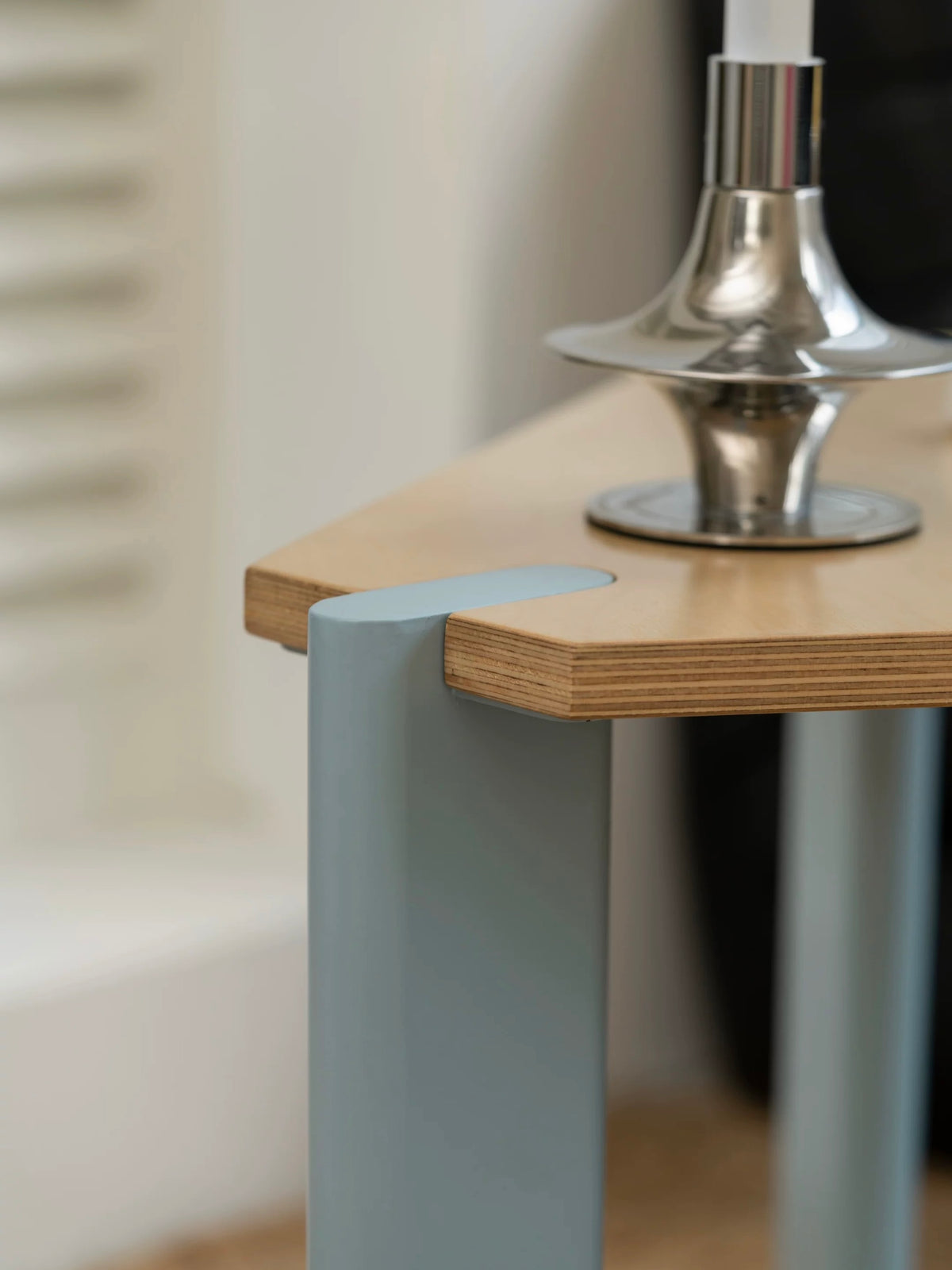Plywood side table close up showing the layered wood edge and a pale metal leg at a clipped corner.