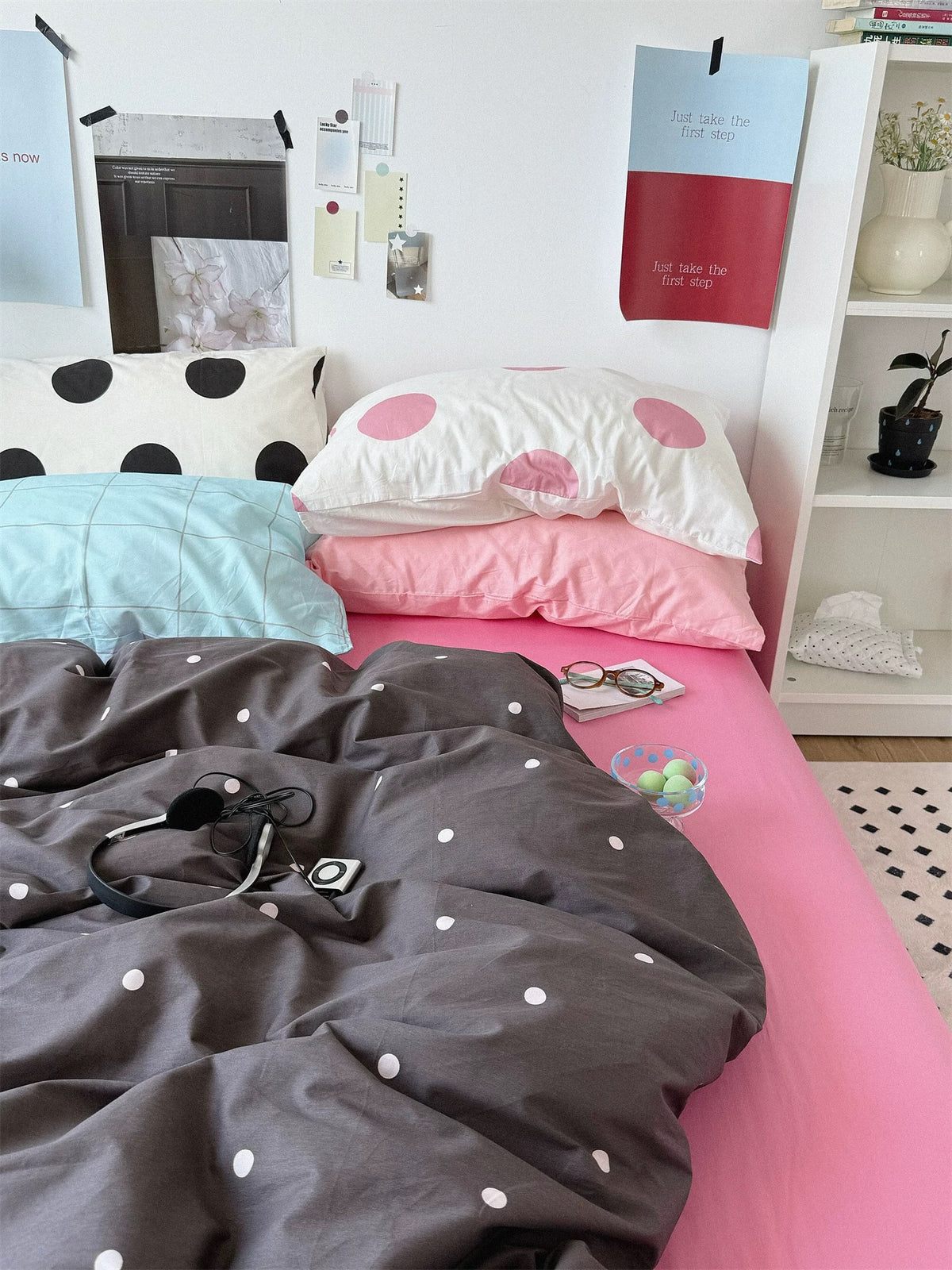 Side view of preppy bedding with contrasting checked and dotted pillows, a plain pink pillow and the dotted cover folded low.