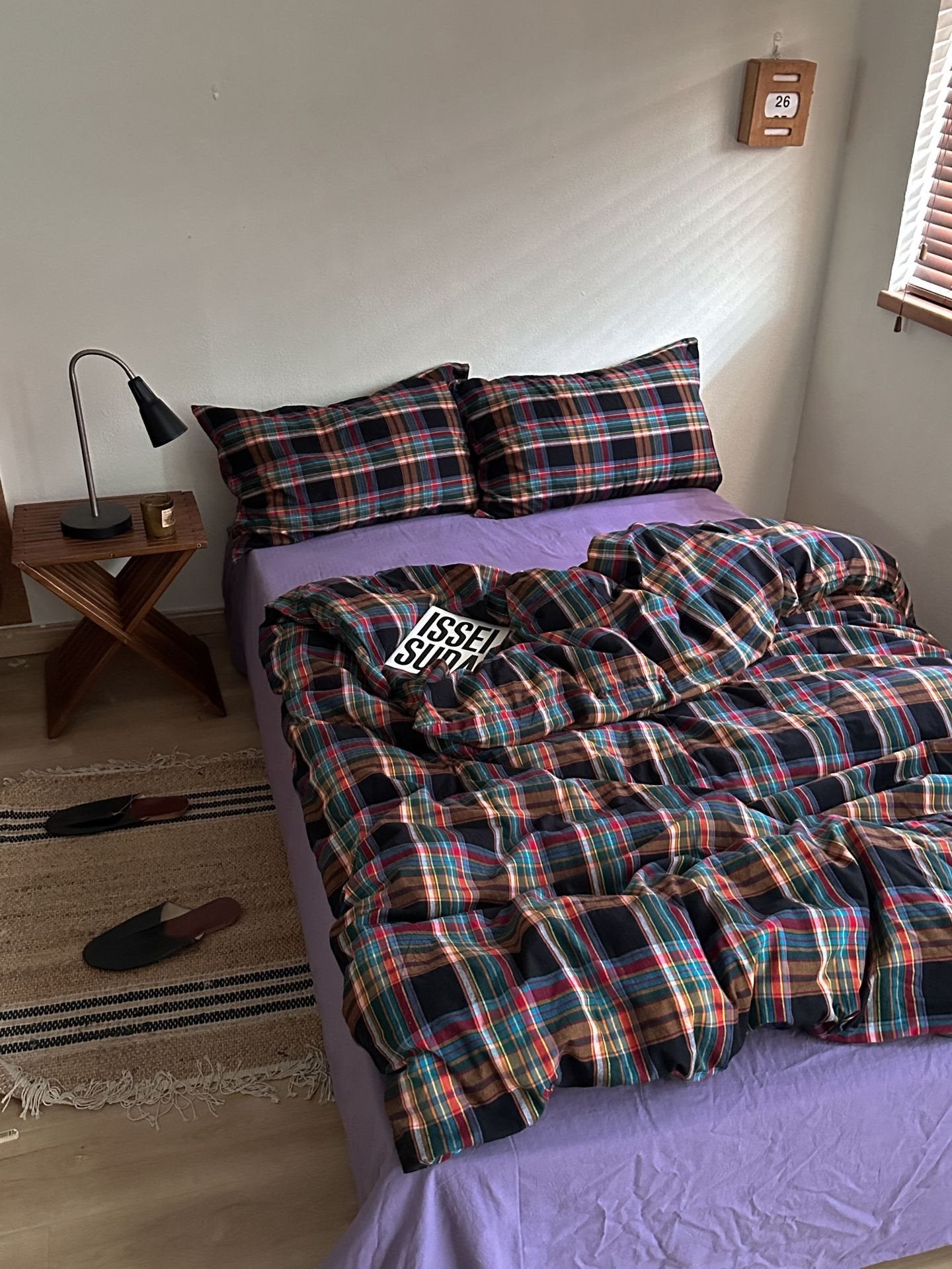 A bed in a bedroom styled with a multicolor plaid bedding set, including a duvet cover and two pillowcases, layered over a solid purple sheet. A small wooden side table with a black lamp sits next to the bed.
