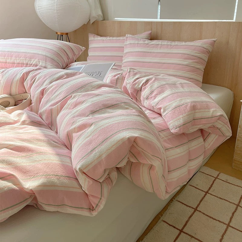 Pink and white striped bedding on a bed with a lamp and window in the background.