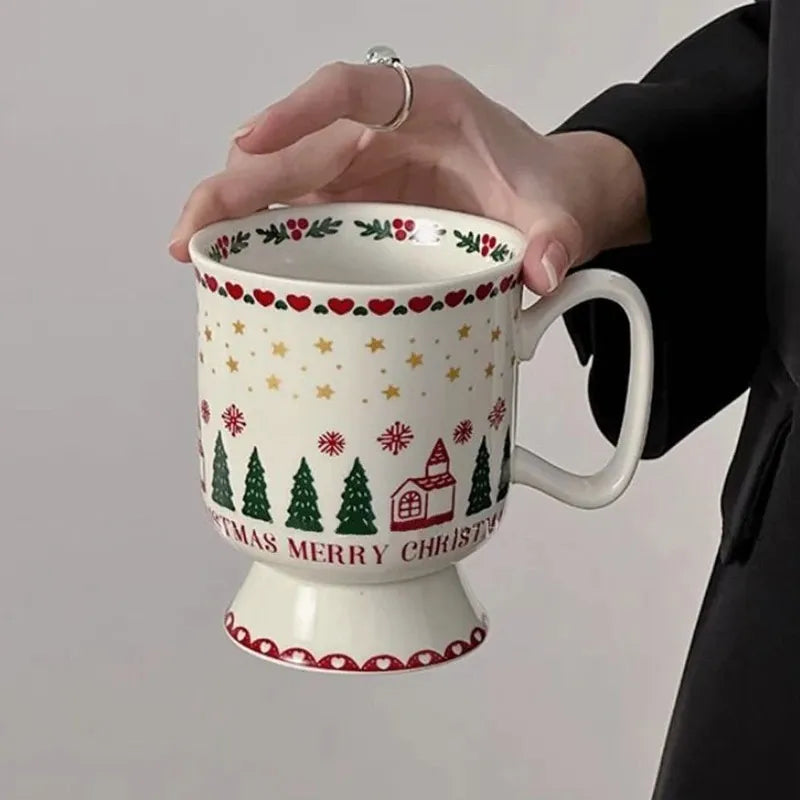 A hand holding a white ceramic Christmas mug with a red and green holiday pattern, featuring pine trees, stars, hearts, and the text 'MERRY CHRISTMAS' on a light gray background.