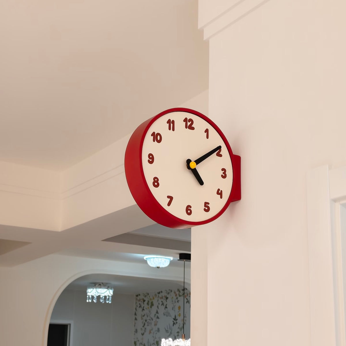 A round wall clock with a red outer frame and a cream-colored face, mounted on a white wall in an indoor setting.