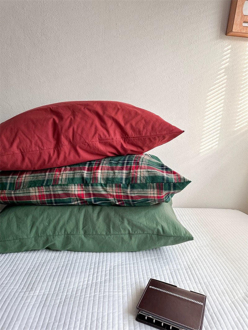 A solid red pillowcase rests above stacked tartan and green pillowcases.