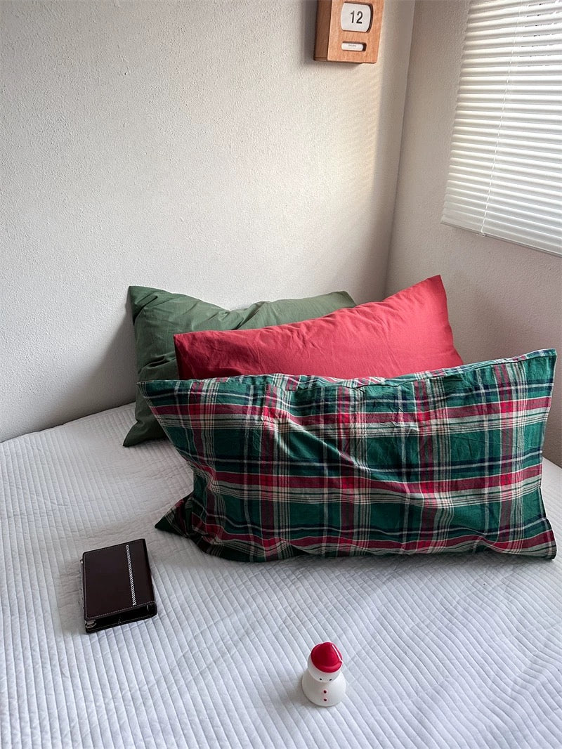 A Christmas pillowcase in green and red tartan sits beside a matching plaid design, with solid red and green pillowcases behind them.