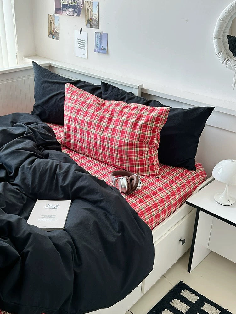 Bed with red plaid blanket and black pillows in a room with white walls and a window.