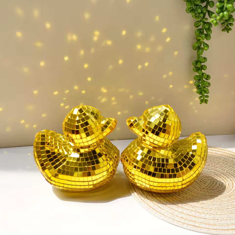 Two gold disco duck decorations on a white surface with a beige wall and green plant in the background.