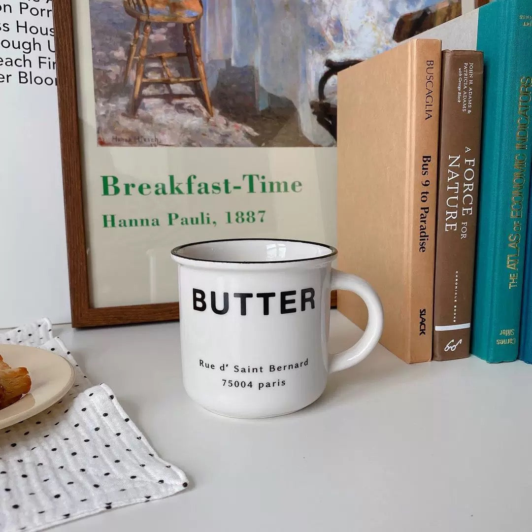 A white ceramic mug with black text reading 'Le Matin & Le Midi Et Le Soir' sits on a blue-and-white patterned saucer with a spoon, next to a baguette and pastry on a bed with white bedding.