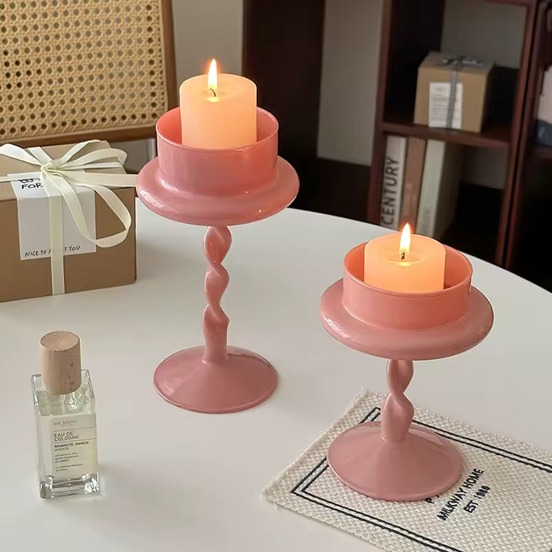 Two pink candle holders with lit candles on a table next to a bottle of perfume.