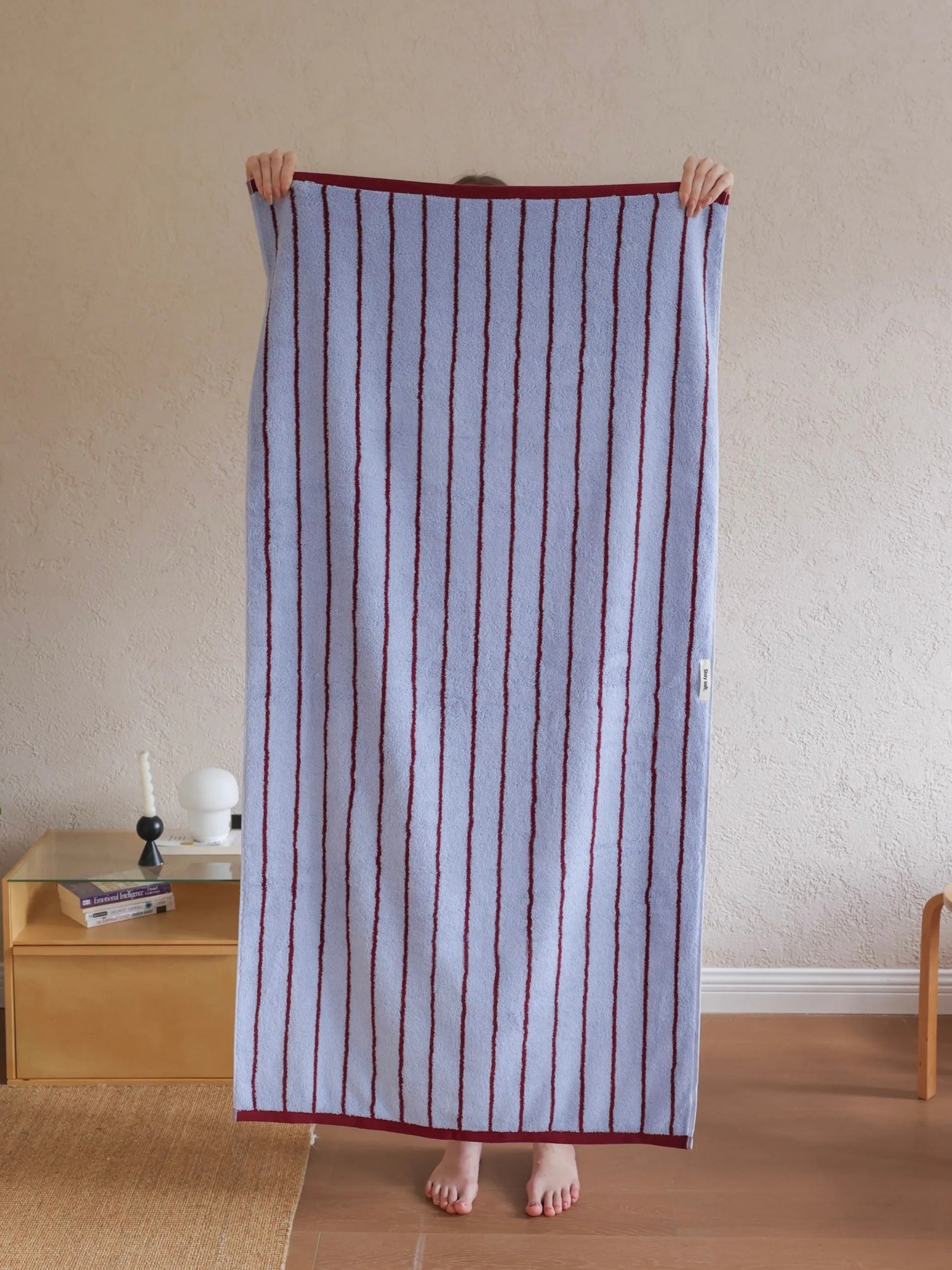 A person holds up a large striped towel or blanket in a home setting. The textile has vertical stripes in dark red/burgundy on a pale lavender-blue background, with a solid dark red border.