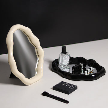 Decorative mirror with a black tray holding a bottle, small box, and other items on a light surface.