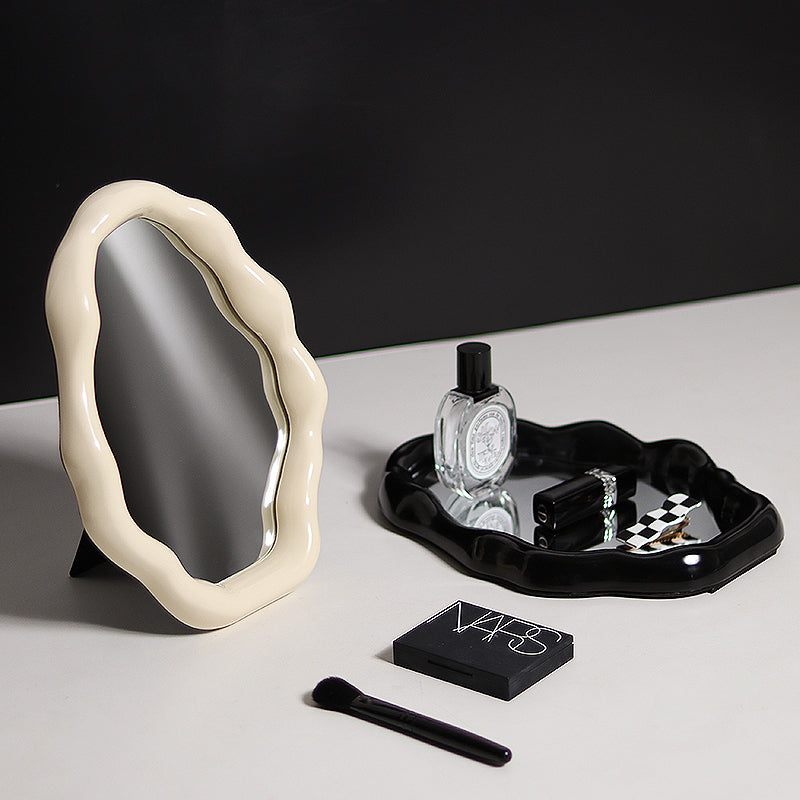 Decorative mirror with a black tray holding a bottle, small box, and other items on a light surface.