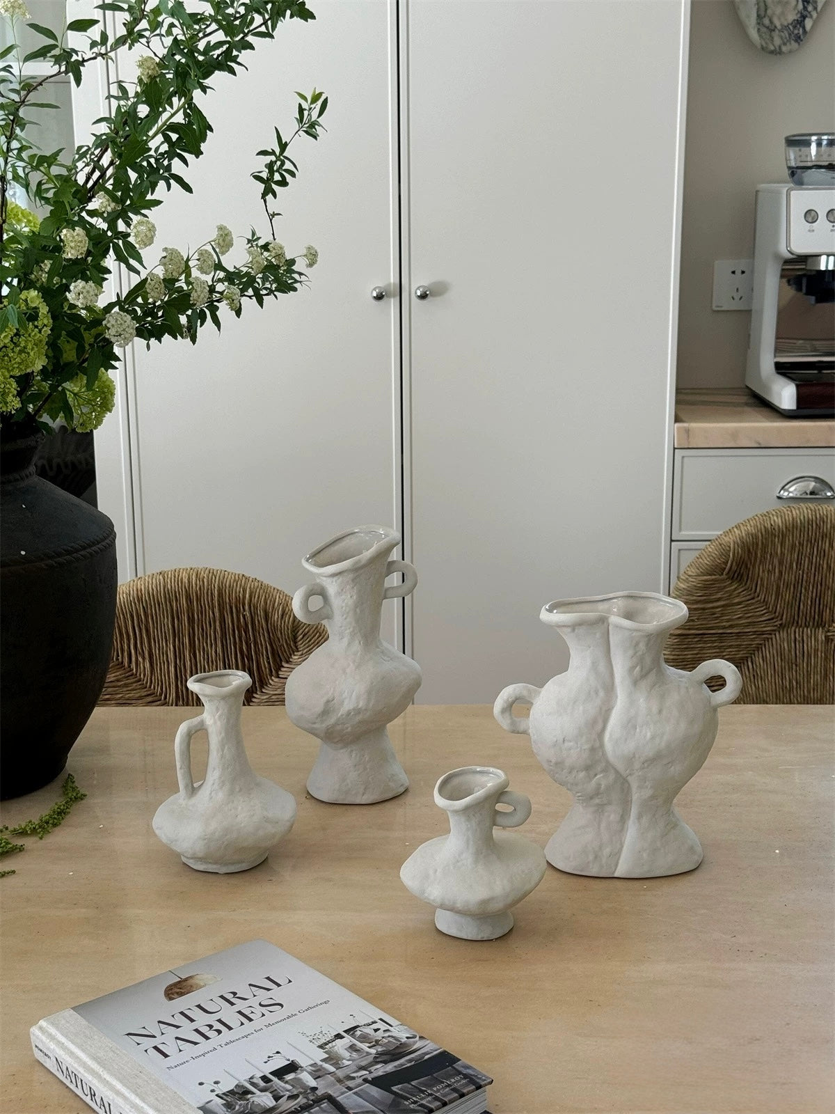 Set of white ceramic vases on a wooden surface with a book titled 'Natural Tables' and a plant in the background.