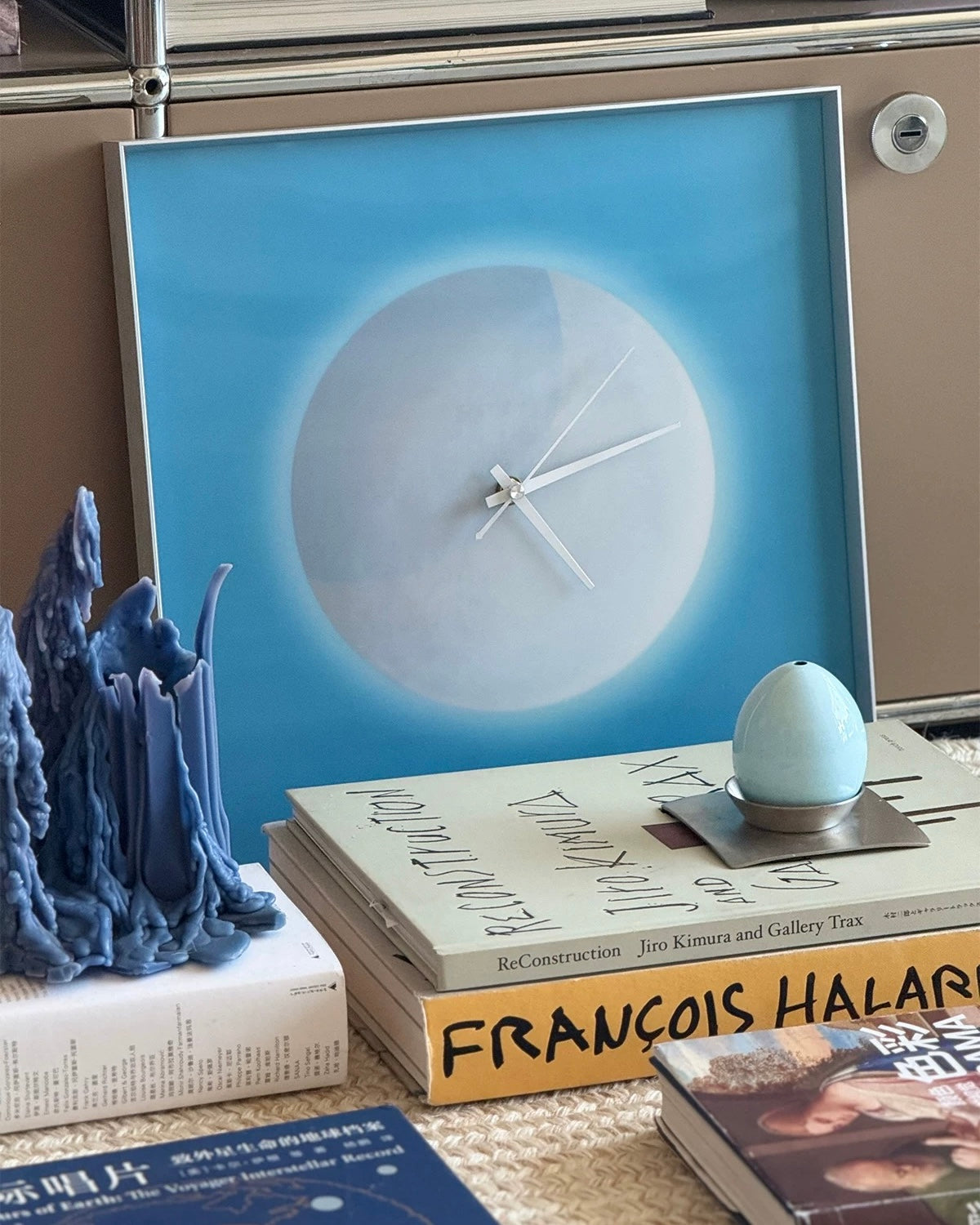 A square wall clock with a blue gradient background and a pale moon face, styled on a tabletop with books, a blue sculptural object, and a small egg-shaped decor piece.