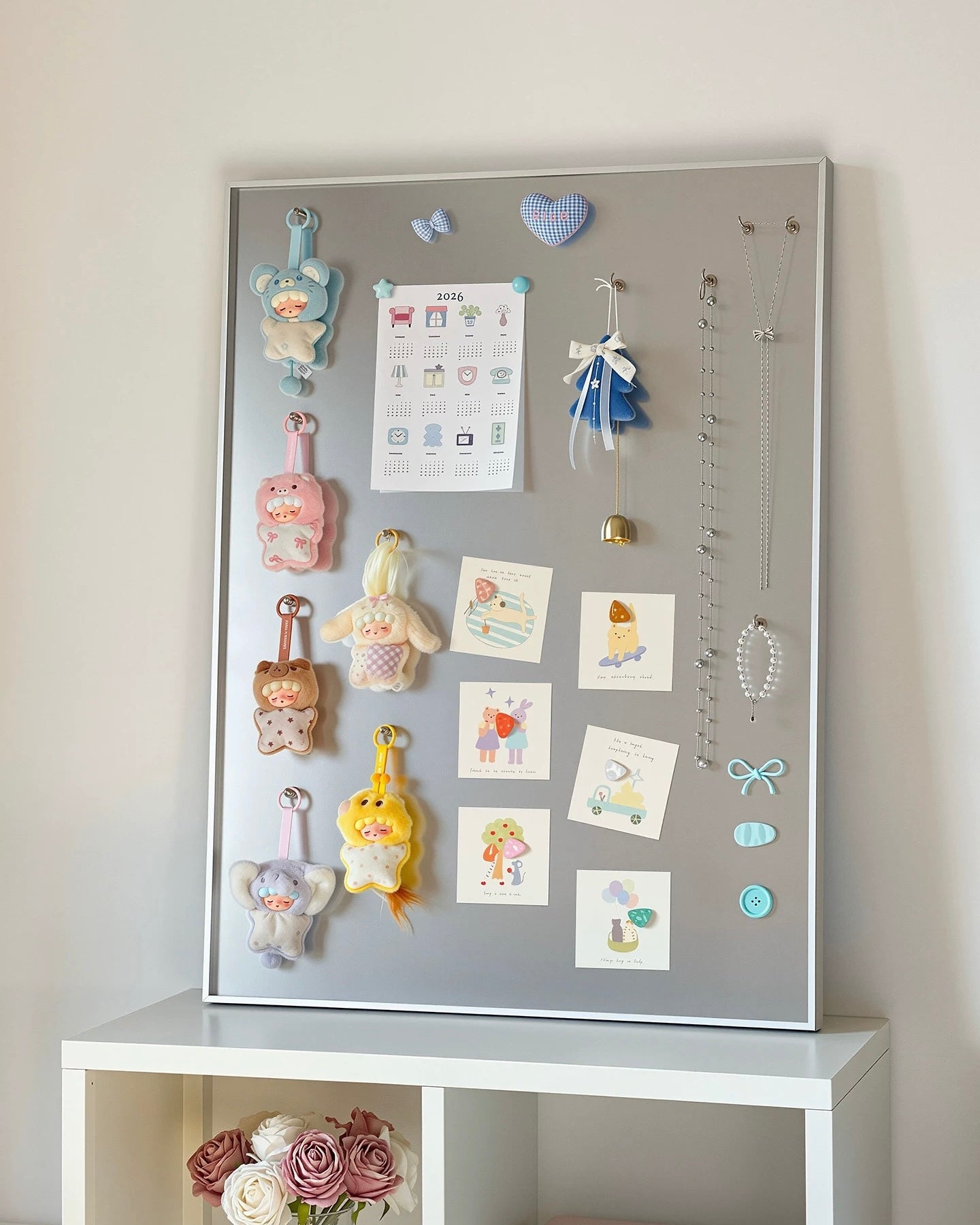 A gray magnetic board mounted on a white wall above a white shelf, decorated with pastel magnets, cards, clips, and hanging accessories.