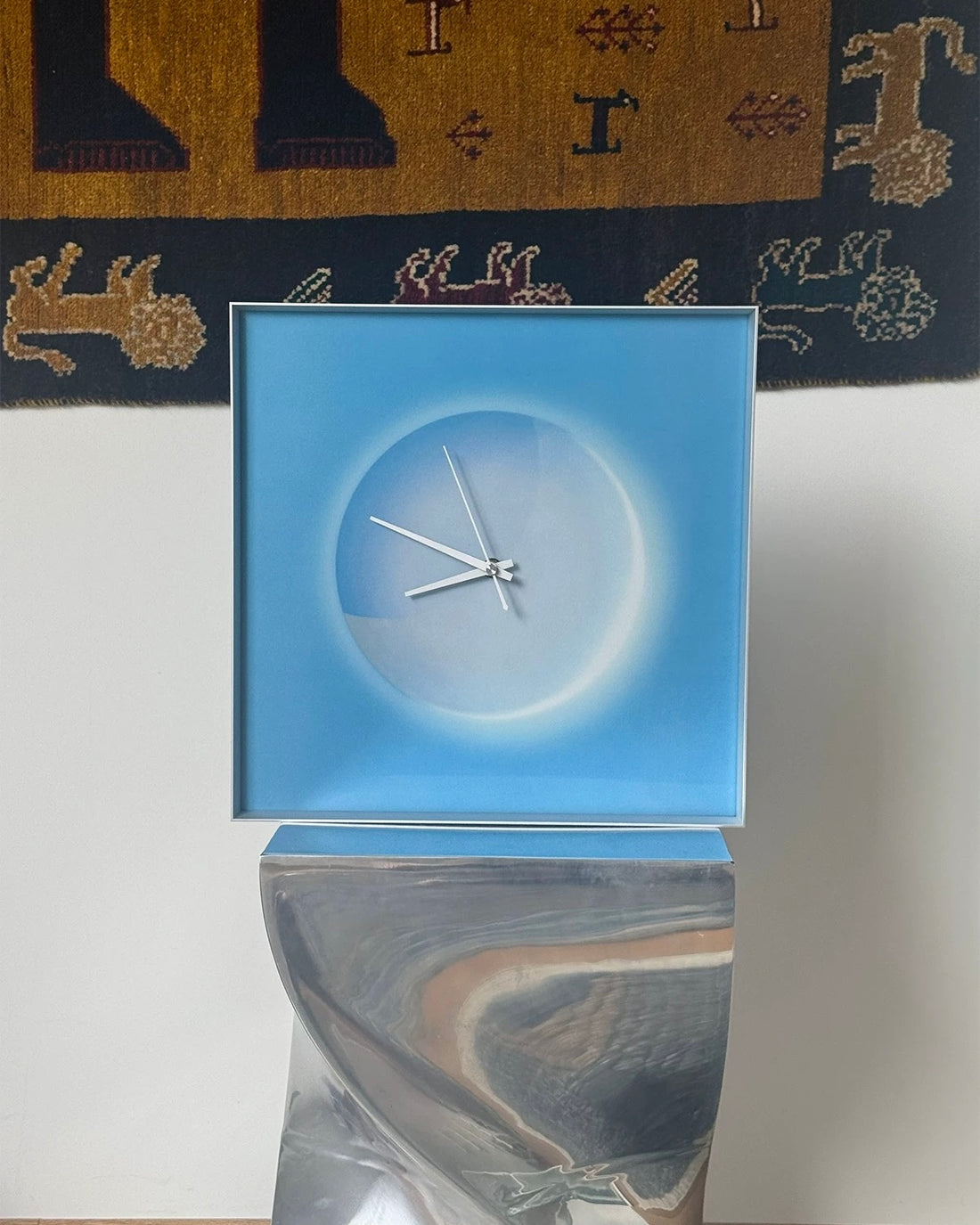 A square framed wall clock with a blue border and a minimalist white circular face sits on a light gray console table in a modern room setting, styled with a candle, decorative boxes, and a chair.