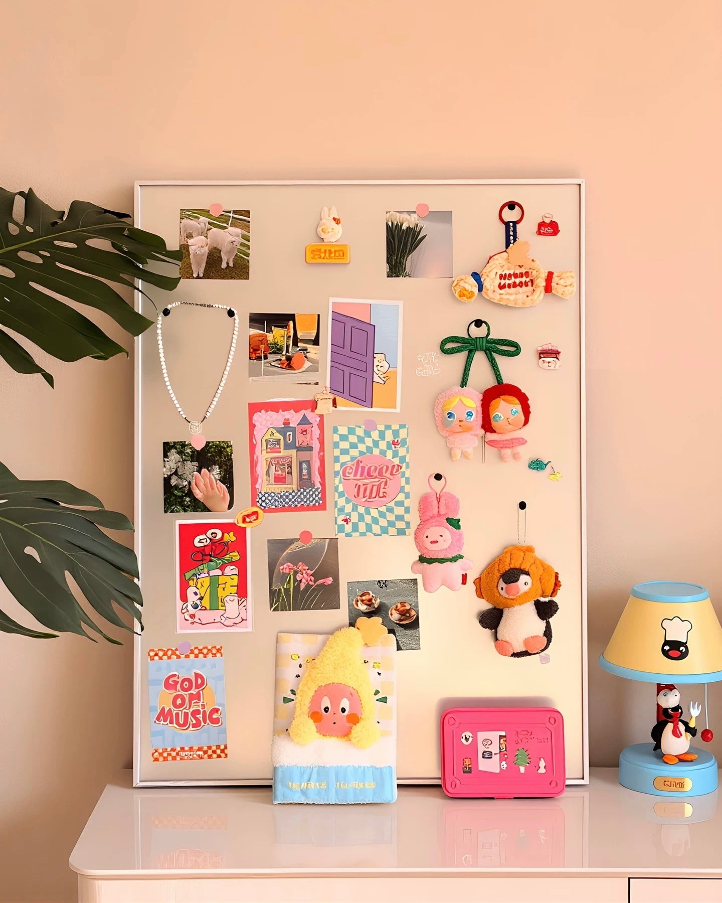 A white magnetic board styled with various cute stationery items, plush toys, and small photos, displayed on a white table against a warm peach-pink wall next to a green plant leaf and a penguin lamp.