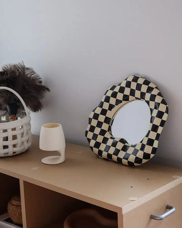 A scalloped-edge oval mirror with a black and cream checkerboard pattern, styled on a light wood cabinet next to a white ceramic vase and a basket with feathers.
