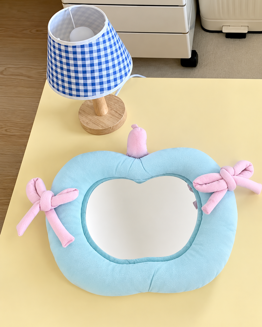 A light blue pumpkin-shaped plush cushion with a mirror in the center and pink bows on the sides, styled on a yellow table next to a blue and white gingham lamp.