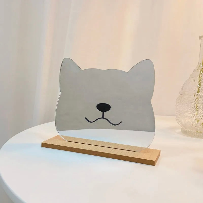 A small decorative tabletop piece shaped like a cat head, made from clear acrylic or glass with a black simple face, mounted on a light wood base. It sits on a white round table with a softly lit neutral background and a blurred glass vase at the side.