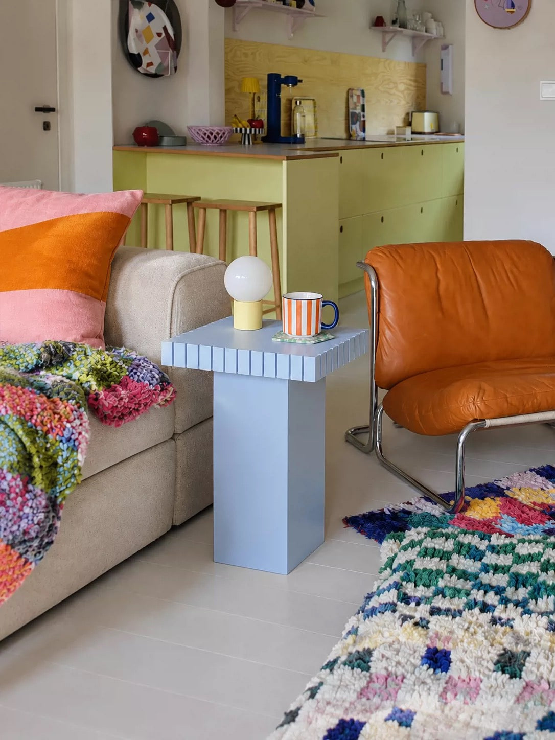 Light blue small side table with a striped mug and a white lamp beside a beige sofa and an orange leather chair
