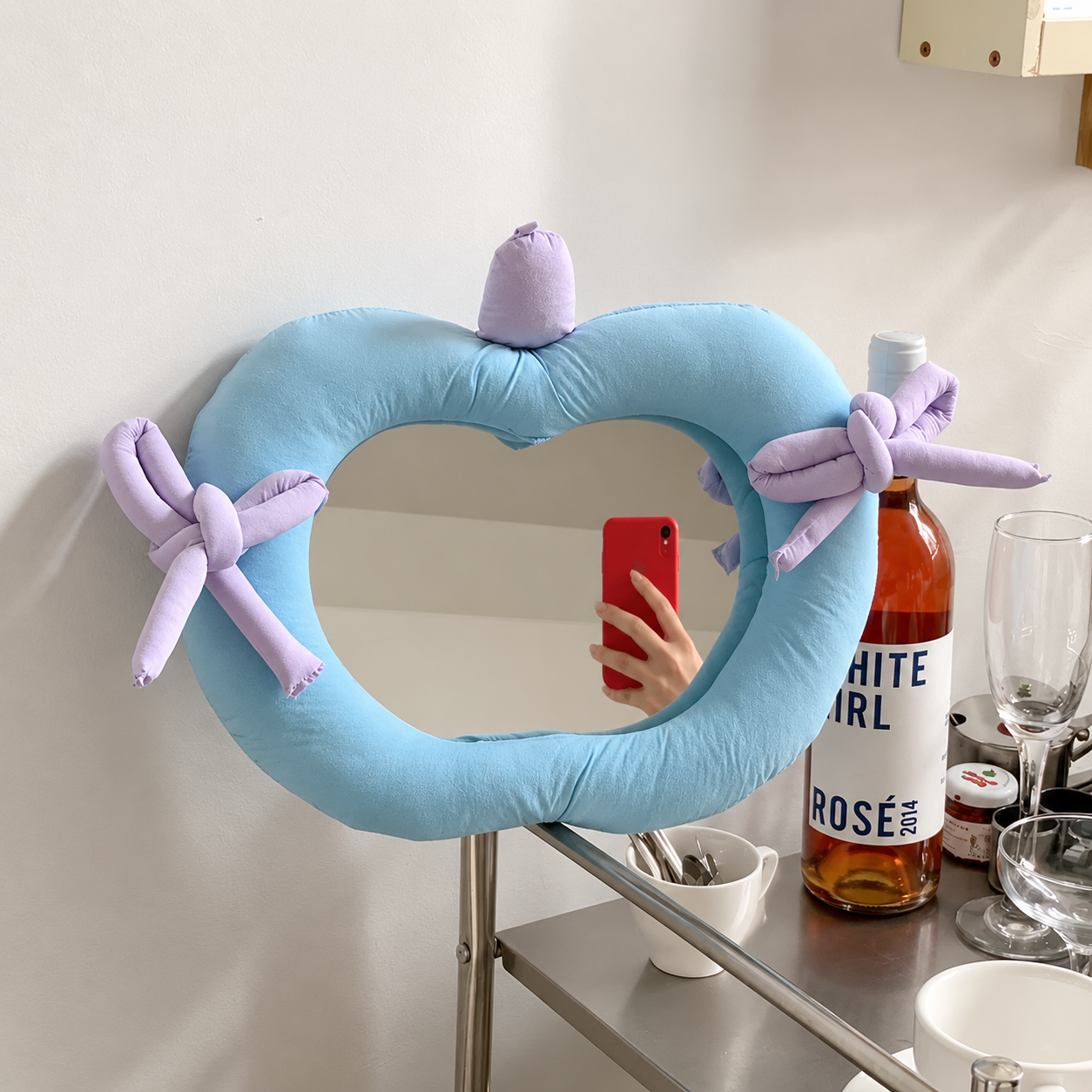 A heart-shaped mirror with a light blue frame and lavender bows, shown in a real-world setting with a person taking a mirror selfie in its reflection.