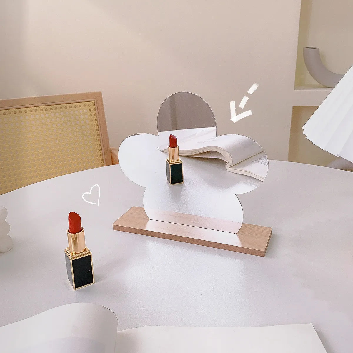A white cloud-shaped tabletop mirror with a wooden base sits on a white table, styled with two lipstick tubes. One lipstick is placed on the tabletop in front of the mirror, and another is positioned on the mirror surface.