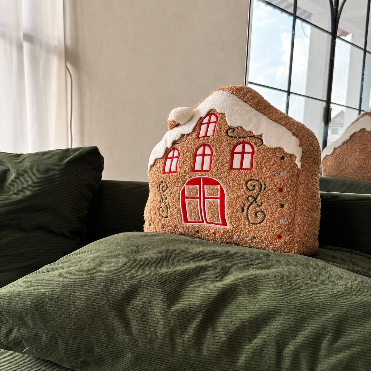 Gingerbread snow house pillow with red windows and snow-covered roof on a green sofa