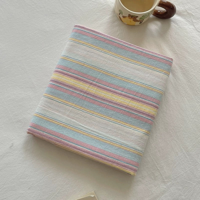 A folded white cotton muslin fabric item, likely a baby blanket or swaddle, featuring pastel horizontal stripes in light blue, pink, yellow, and purple. It is styled on a white textured fabric surface next to a ceramic mug.