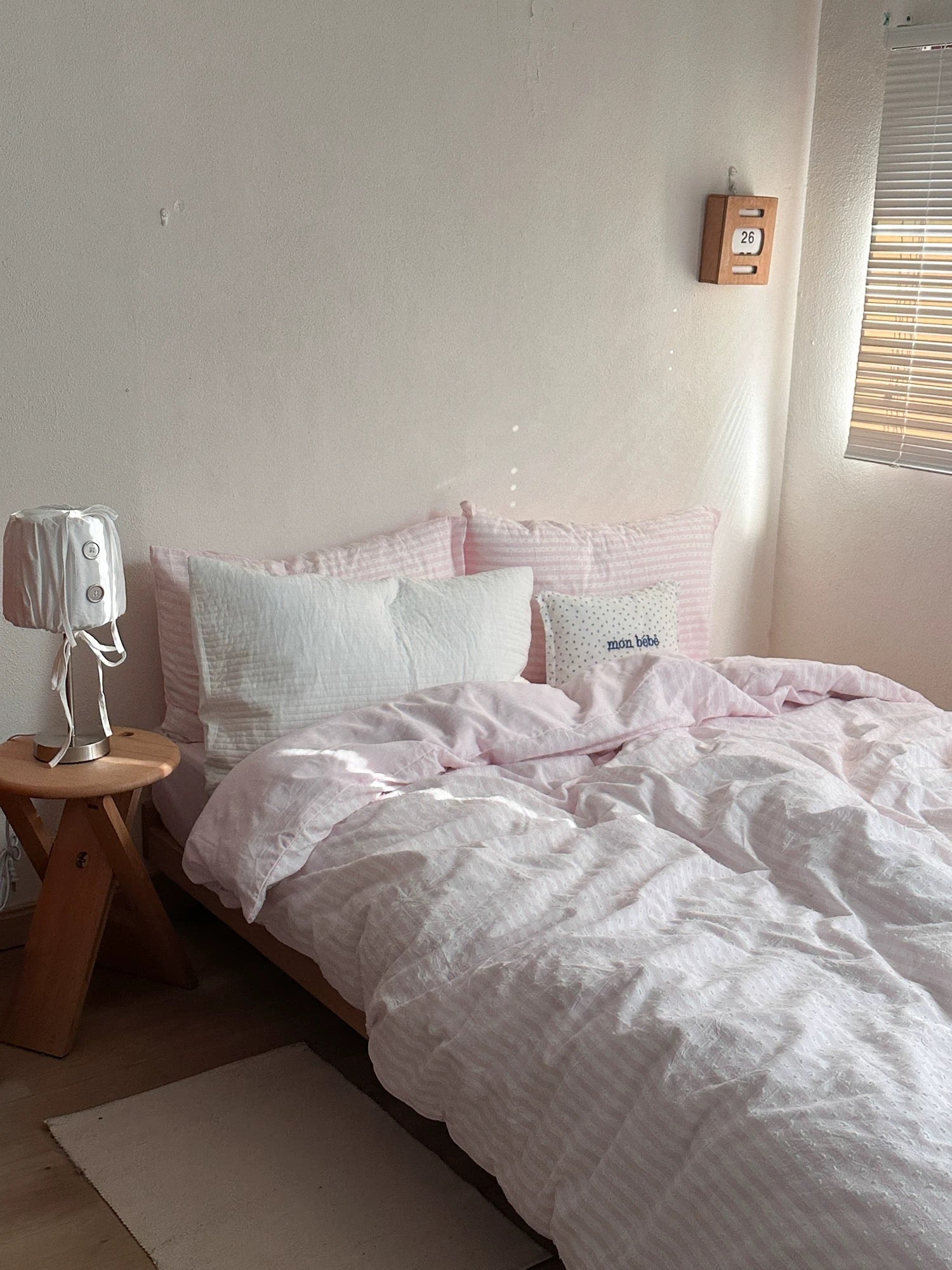 A bed dressed in light pink and white striped bedding, including a duvet cover and matching pillows, styled in a minimalist bedroom setting with a small wooden side table and a white lamp.
