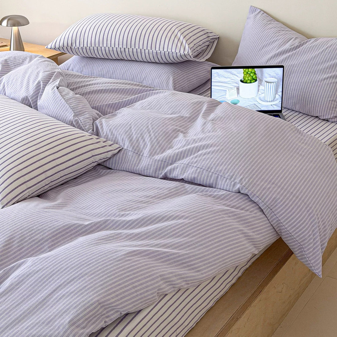 A minimalist platform bed with light wood flooring, dressed in a lavender and white striped bedding set with matching pillows, a small wooden bedside table, and a laptop on the bed.