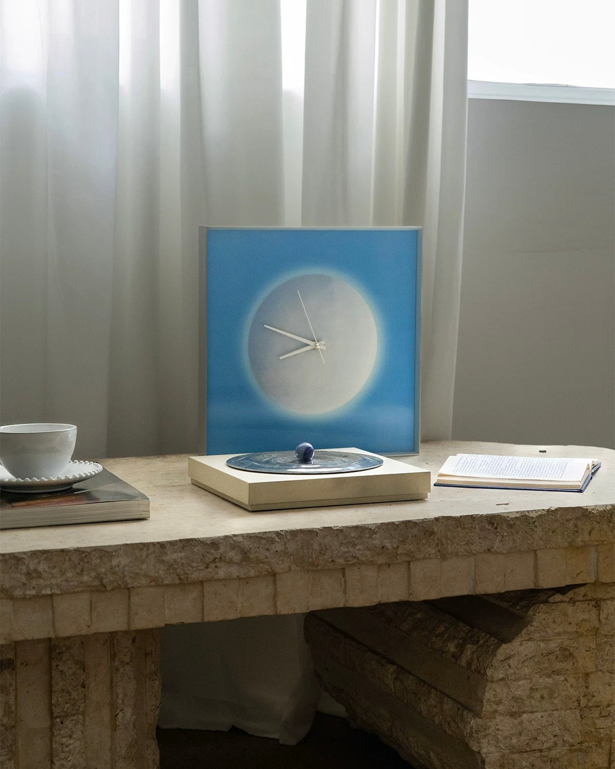 A square wall clock with a blue gradient background and a white circular clock face, styled on a stone table next to a cup, notebook, and other decor items in a bright room.