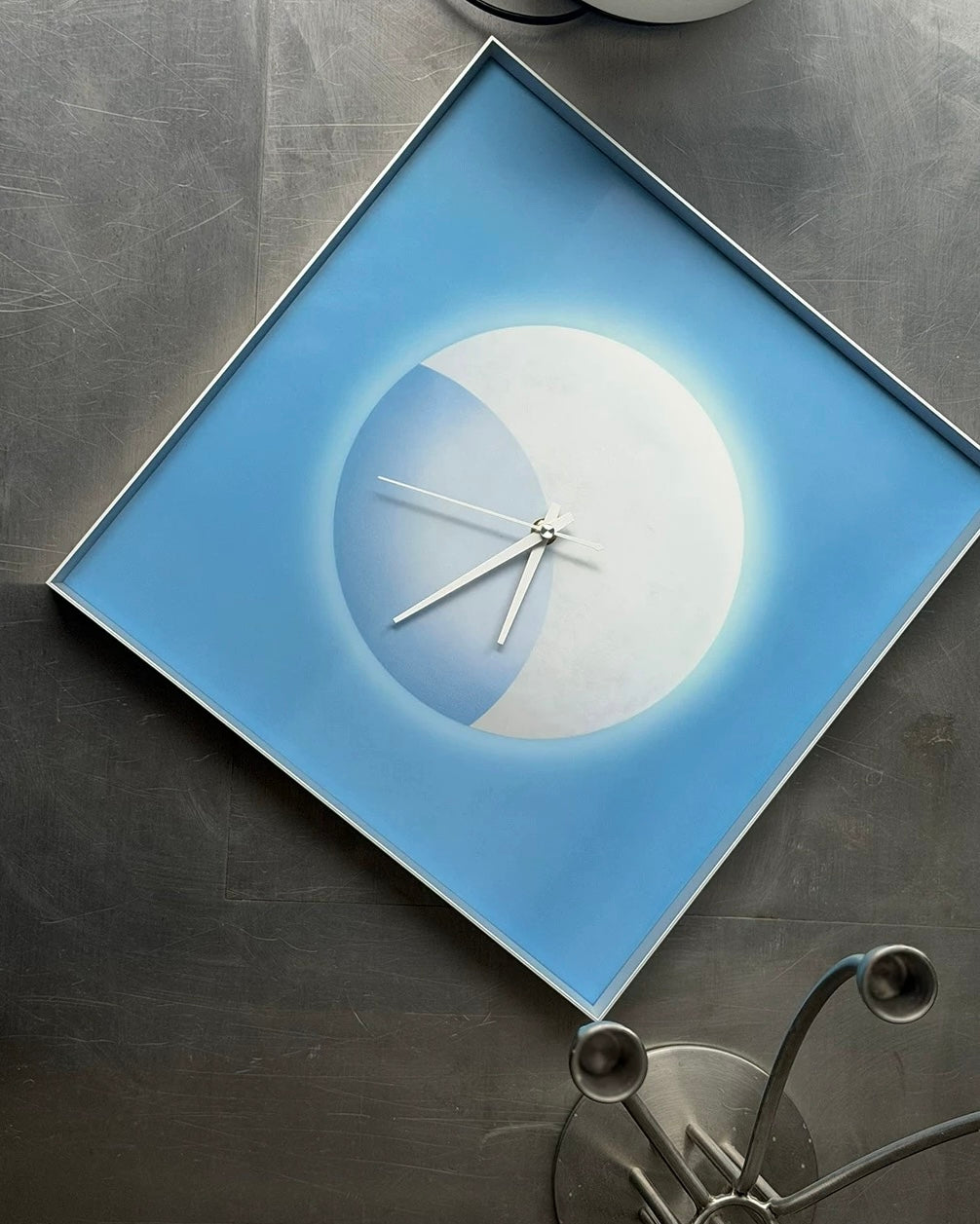 A square wall clock with a light blue frame and a minimalist moon design in white and light blue, mounted on a dark textured wall.