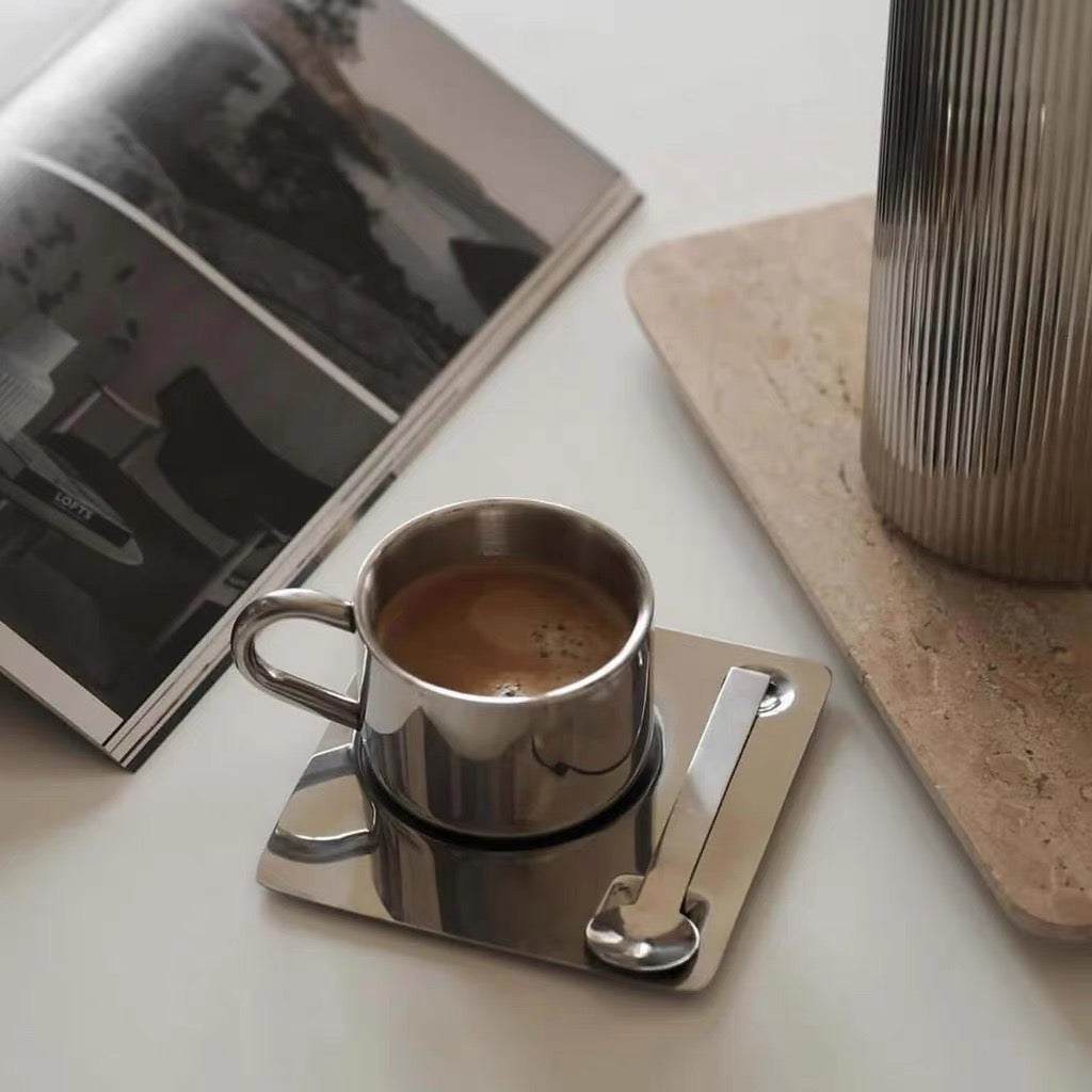 Silver coffee cup with saucer on a white surface next to a book and cylindrical container.