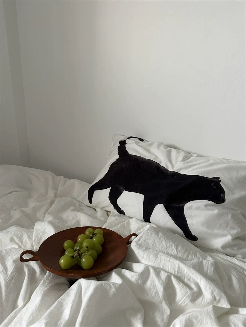 Black cat on a white pillow with a plate of grapes on a white bed.