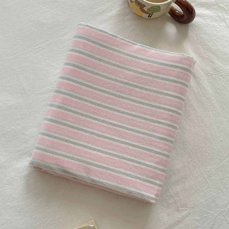 A folded square fabric item, likely a pouch or small bag, with horizontal pink and light gray stripes, placed on a white textured fabric surface next to a small decorative ceramic cup.