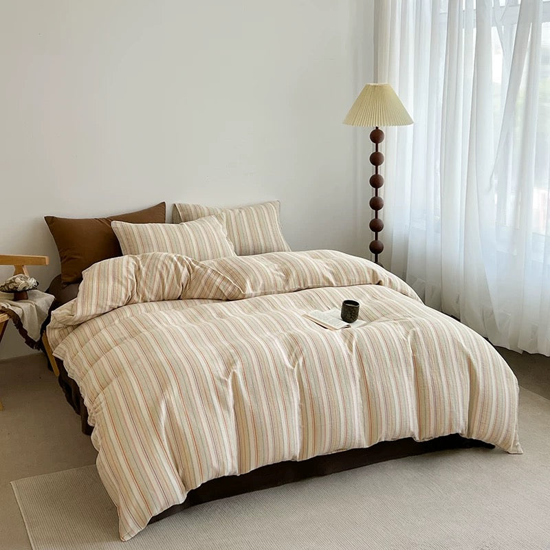 A bed dressed in a beige and cream striped duvet cover and matching pillow shams, styled in a bright bedroom with white curtains, a floor lamp, and a small side table.