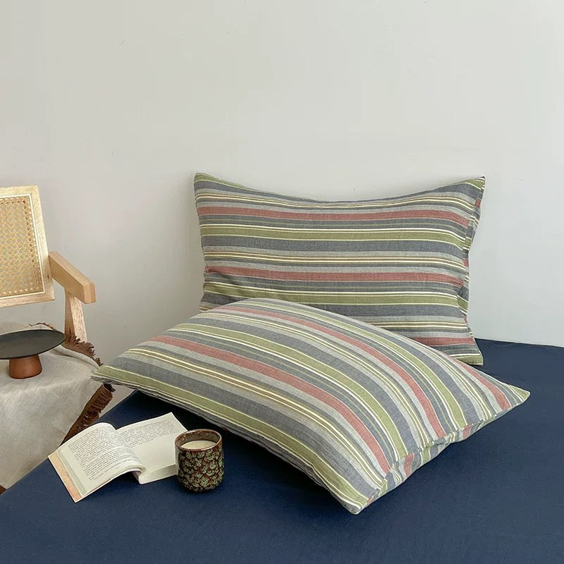 Two striped cotton pillowcases in muted earthy tones of gray, green, pink, and beige, styled on a navy blue bed with a book and candle nearby.