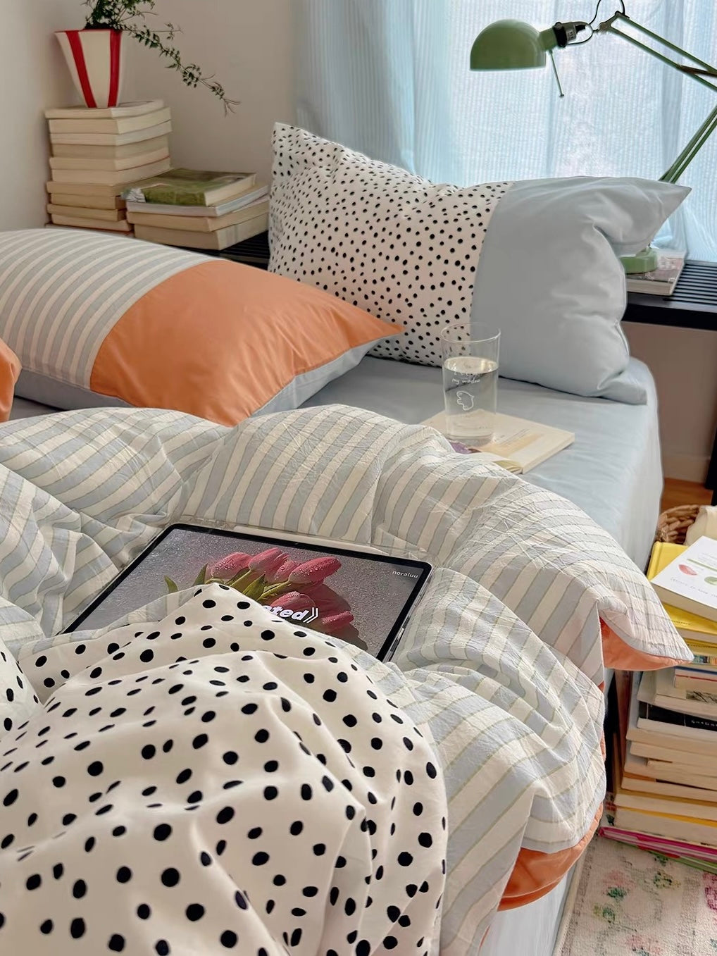 Striped and polka dot bedding is shown beside a stack of books, with a glass and book on the blue sheet.