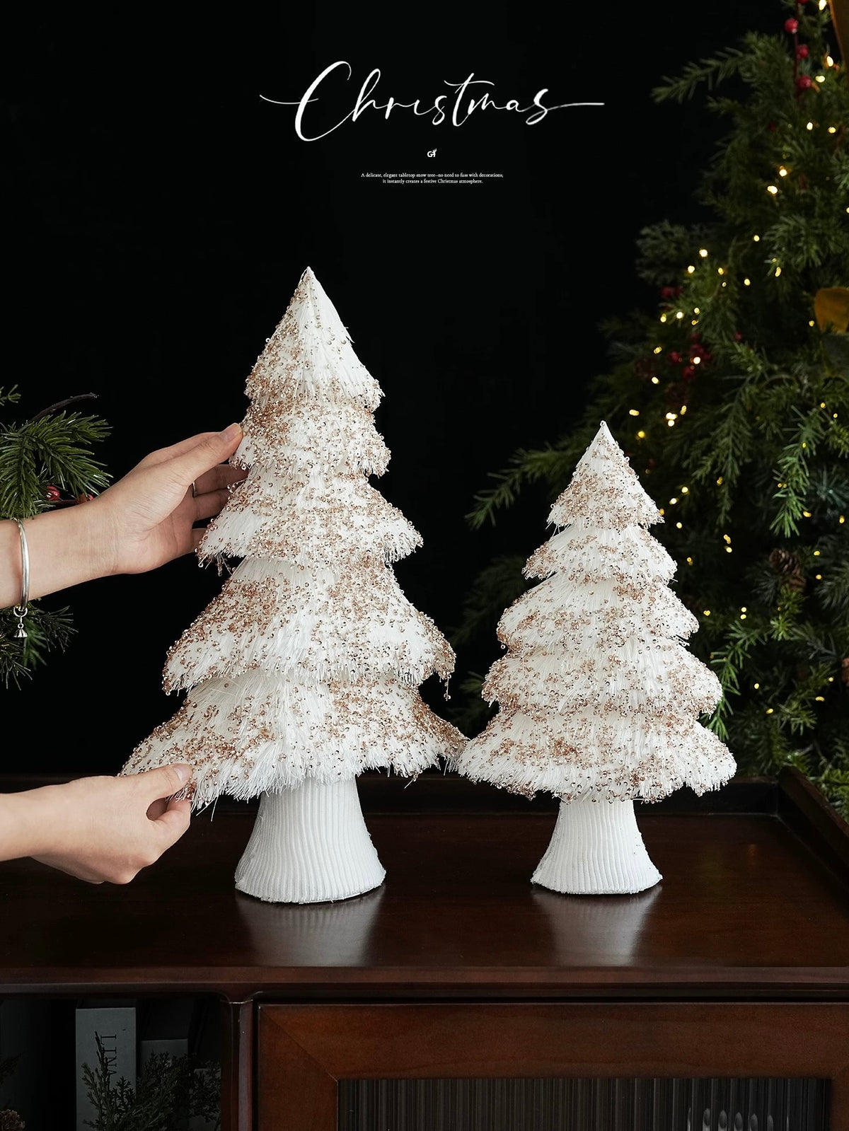 A tiered Christmas tree is shown beside another tree while a hand reaches toward its white fringed branches.