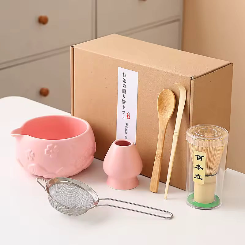 Tea-making set with pink bowl, small vase, wooden spoon, and other tools in front of a cardboard box.