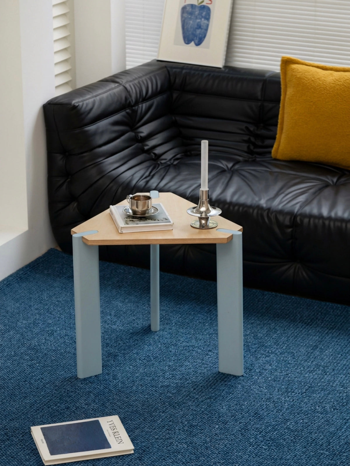Triangular side table shown at an angle with pale metal legs and a natural wood top beside a black sofa.