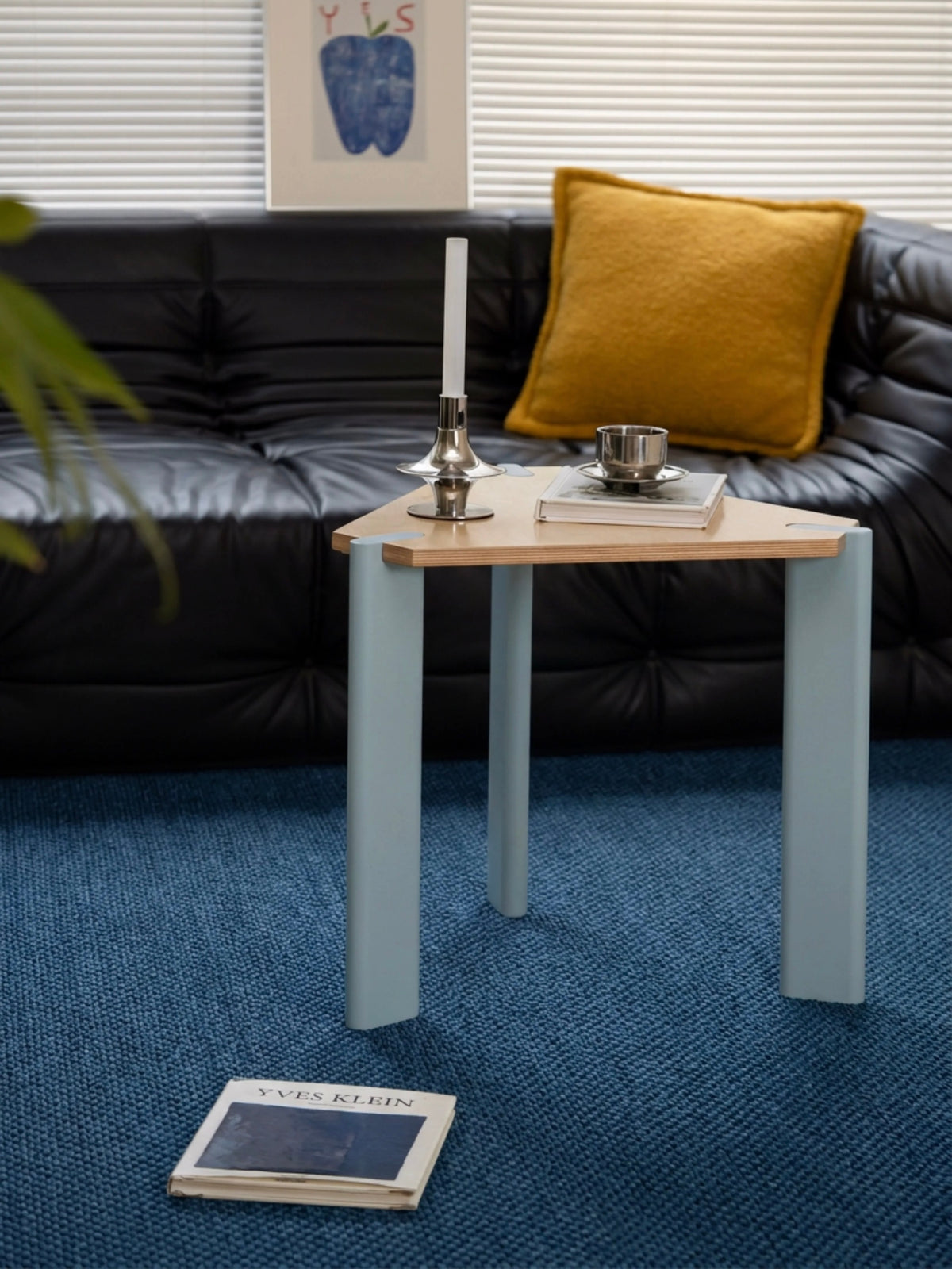 Triangular side table with a natural wood top and pale metal legs beside a black sofa and yellow pillow.