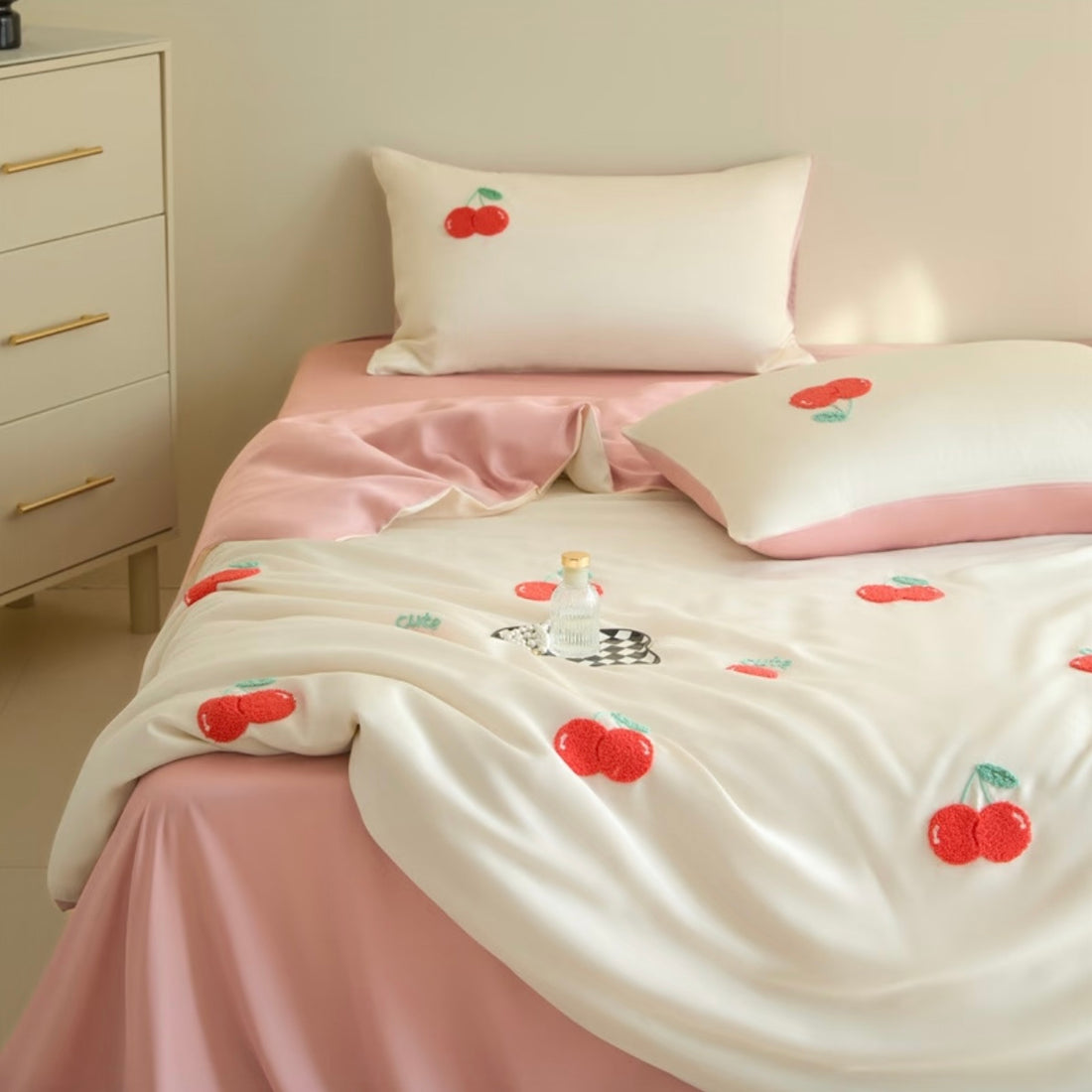 A neatly made bed in a softly lit bedroom, featuring white bedding with embroidered red cherries and a pink satin sheet and pillowcases. A small bedside table with a glowing lamp is visible on the right.